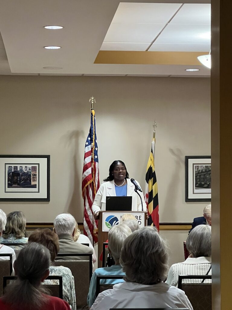Esther Wells speaking at a podium beside the United States and Maryland flags